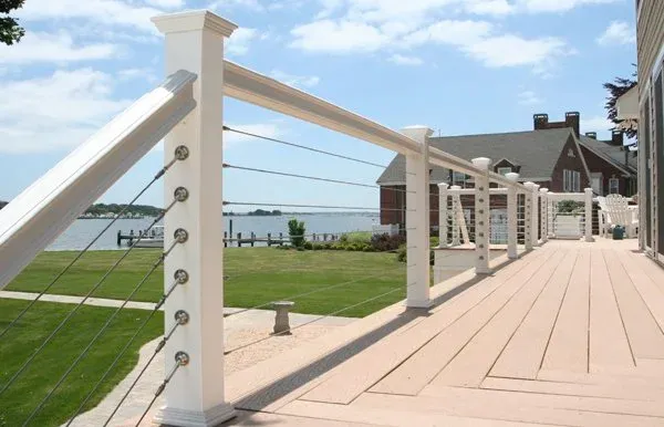 A spacious wooden deck with white railing overlooking a lake, with a house nearby, under a partly cloudy sky.