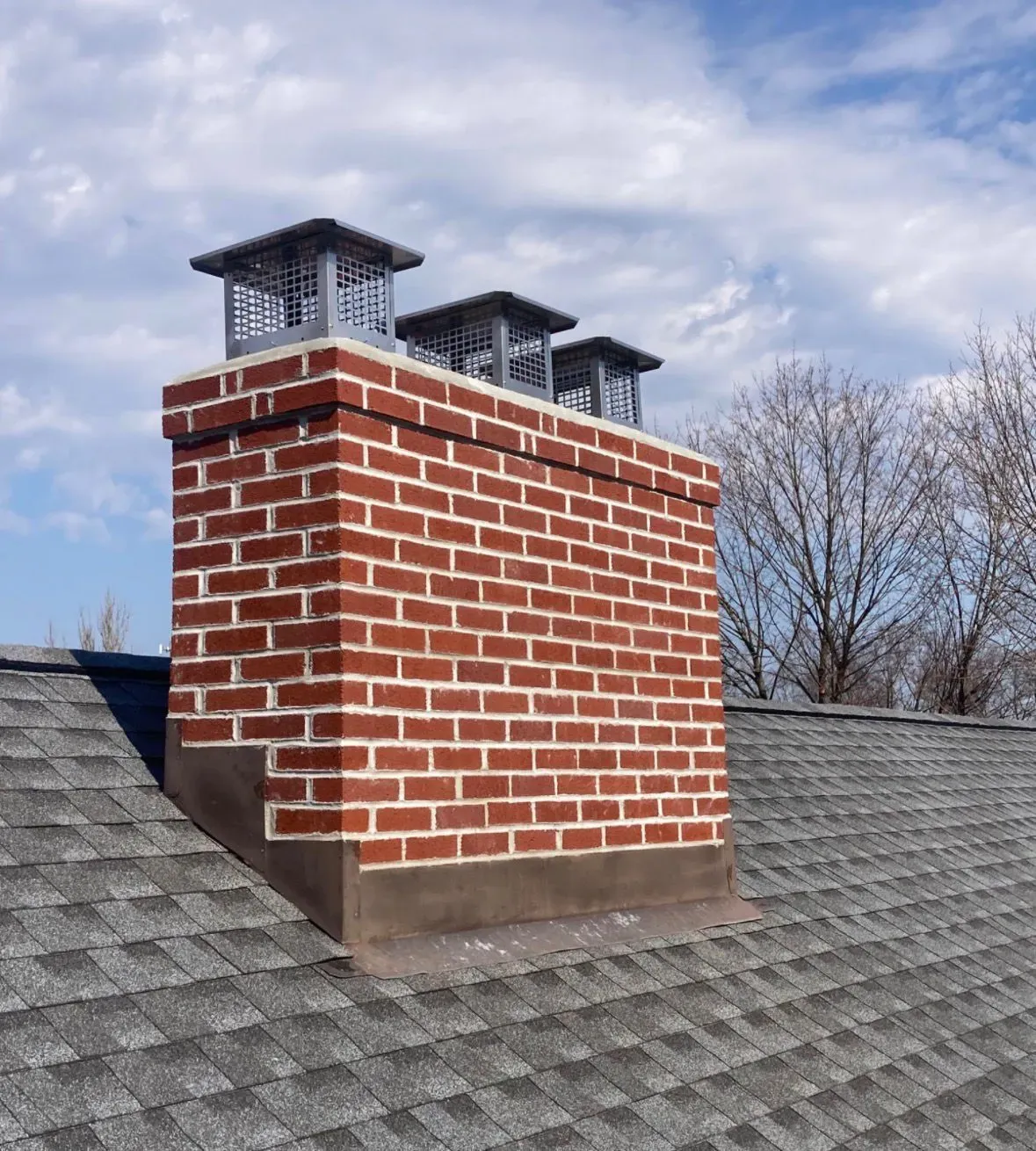 Brick chimney with metal caps on a gray shingled roof under a cloudy sky