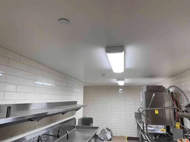 Commercial kitchen with stainless steel appliances, white tile walls, and fluorescent lighting.
