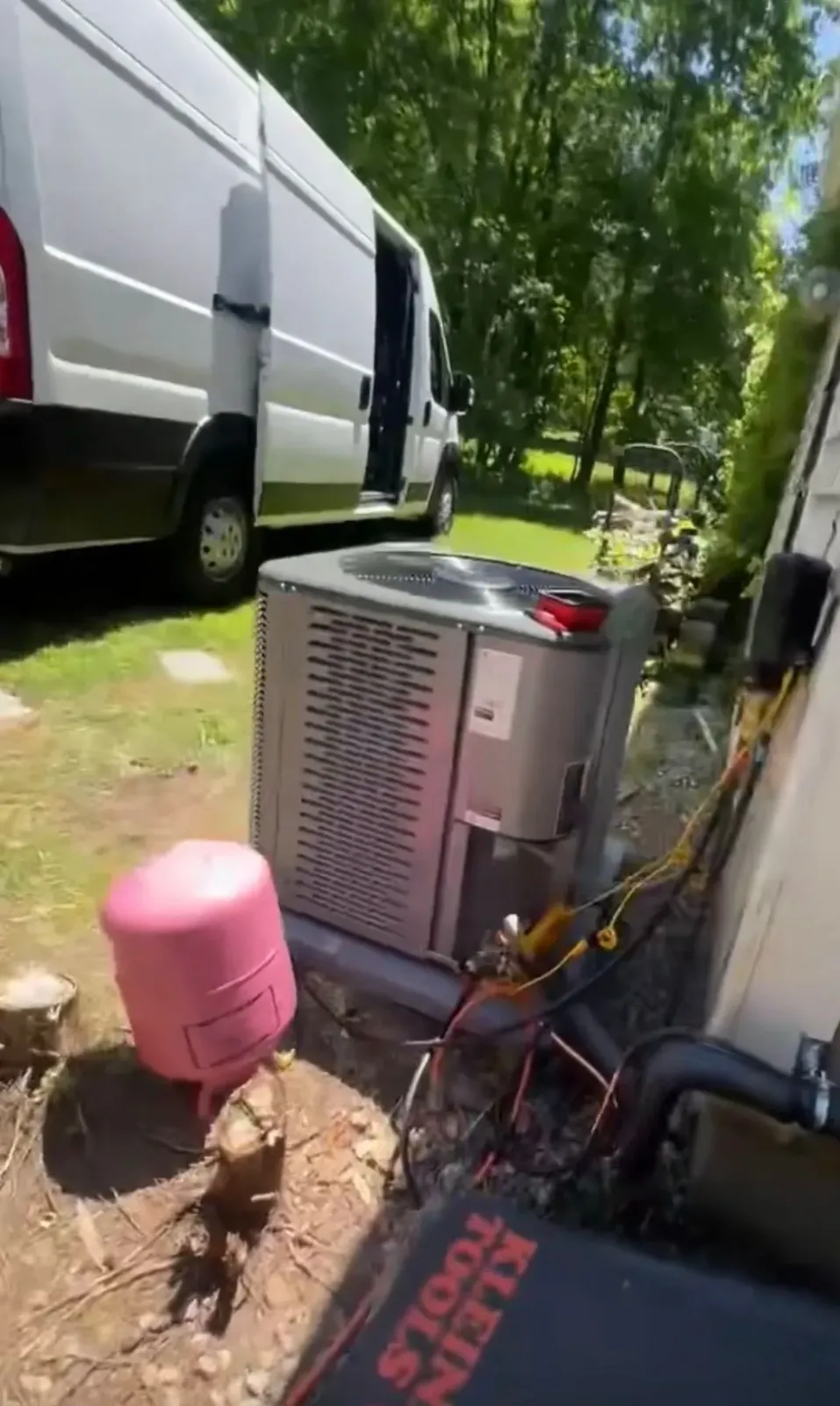 Outdoor utility area with an RV, gray HVAC unit, pink tank, and hoses beside a wall and trees.