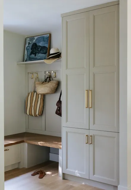 A corner of a room with a white built-in cabinet, gold handles, and a bench with a pair of brown sandals on the floor. Above the bench, a wall has gold hooks holding a striped bag, a hat, and a purse. A shelf above holds a painting, a white hat, and a hat with a wide brim.