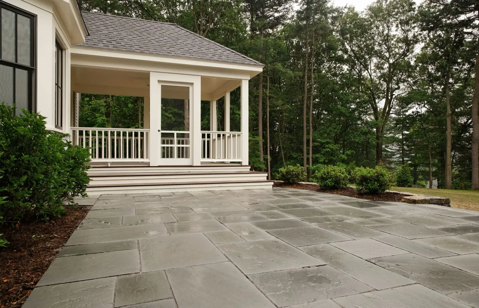 Covered porch beside large stone patio in a wooded backyard