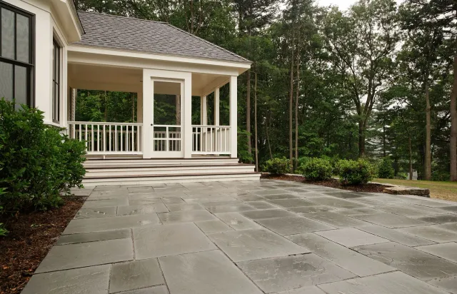 Covered porch beside large stone patio in a wooded backyard