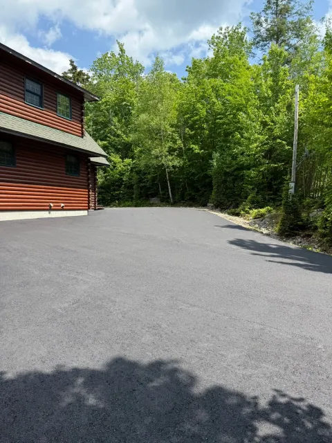 A log cabin with a dark metal roof sits beside a wide, freshly paved gravel driveway, framed by dense green trees.