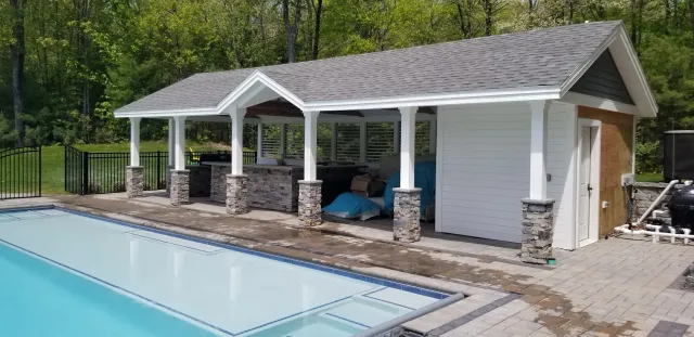 Backyard pool area with a covered outdoor kitchen and storage shed, surrounded by a forested area with green trees.