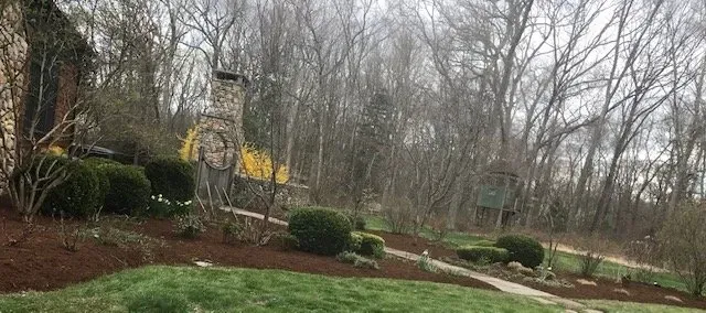 Stone chimney, garden path and shrubs, trees in the background. Overcast sky.