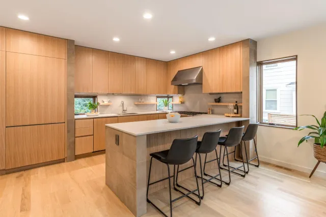 Modern kitchen with wooden cabinets, a central island with a white countertop, four black barstools, and a window between two shelves.