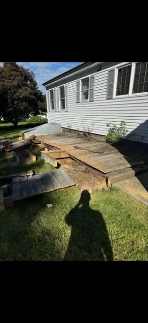 A person's shadow falls on a grassy yard next to a white house with a partially dismantled wooden deck.