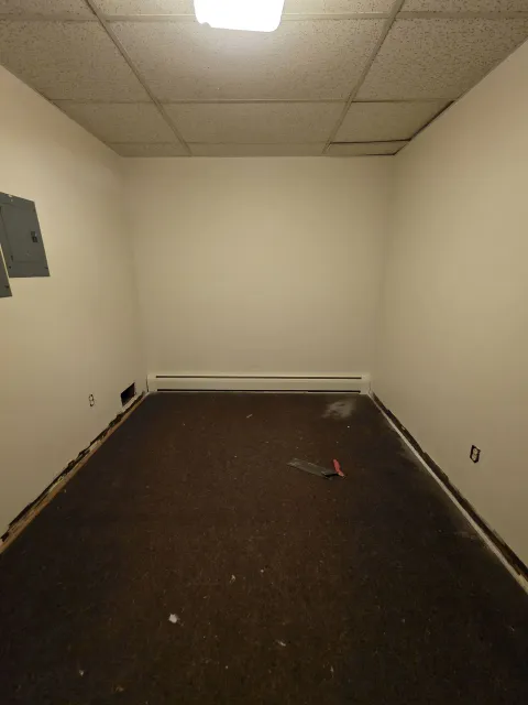 Empty beige room with drop ceiling, dark carpet, and wall utility panels.