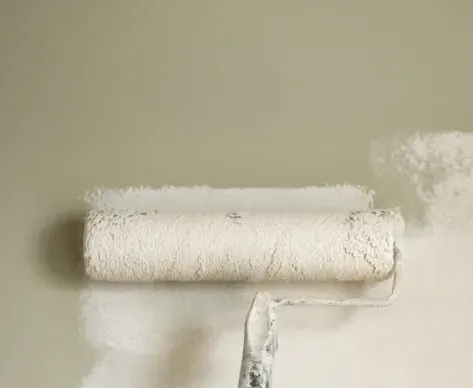 Paint roller applying white paint to a beige wall, leaving a fresh coated strip.
