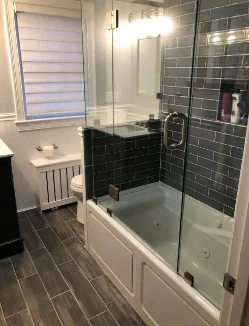 Modern bathroom with a glass shower enclosure, dark tiled walls, and white tub.