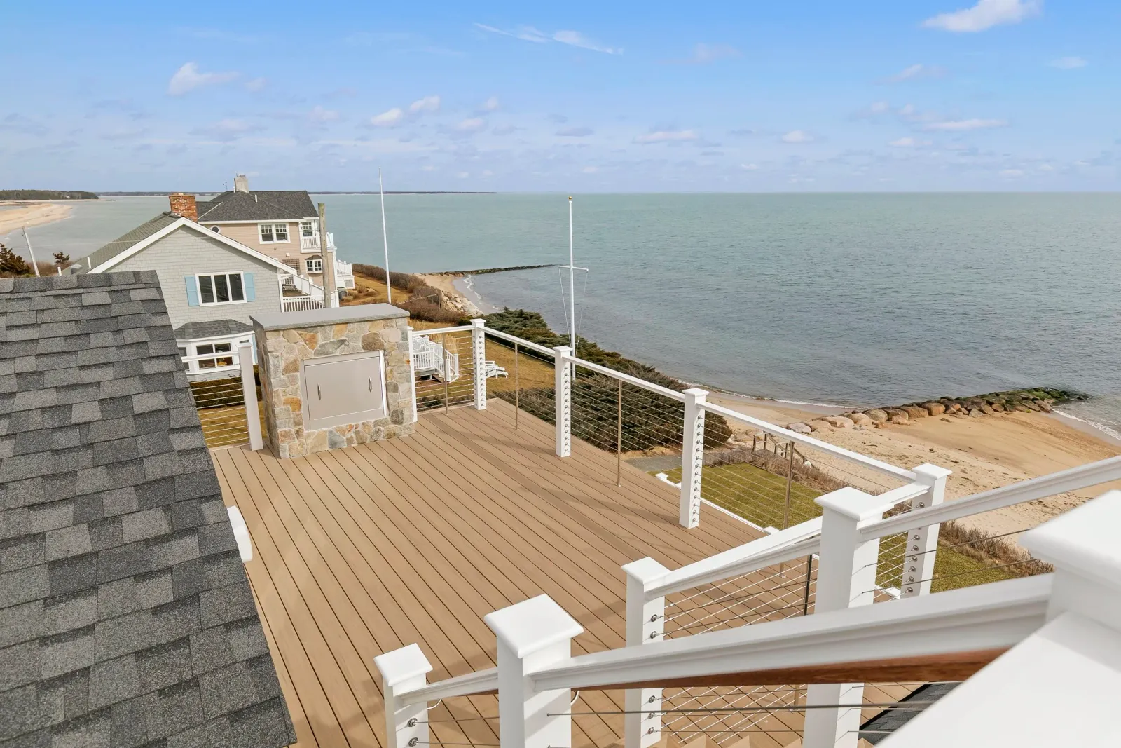 Overlooking a coastal backyard deck with a view of the ocean and sandy beach with rocks, neighboring houses, some beach and water scenery, blue sky with scattered clouds.