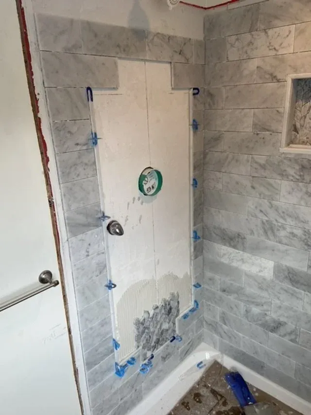 Bathroom wall showing a partially tiled area with a toilet behind a cardboard cover, blue painter's tape around the tiles, and construction materials on the floor.