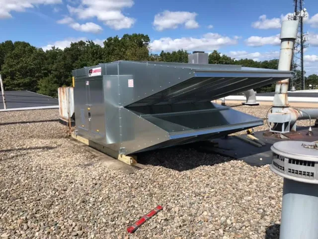 Industrial rooftop HVAC unit on gravel surface with trees and blue sky in background.