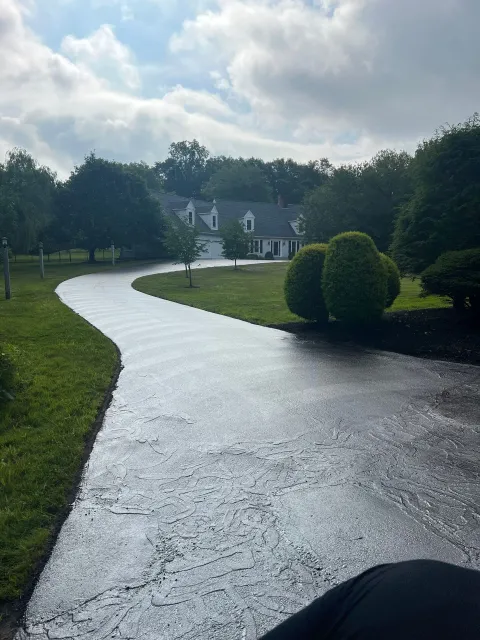 Long, winding driveway leading to a white house, surrounded by green trees and grass, under a cloudy sky.