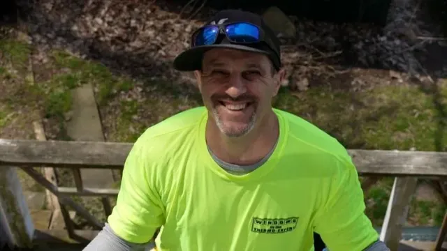 Smiling man in neon yellow shirt and black cap sitting outdoors by a wooden fence