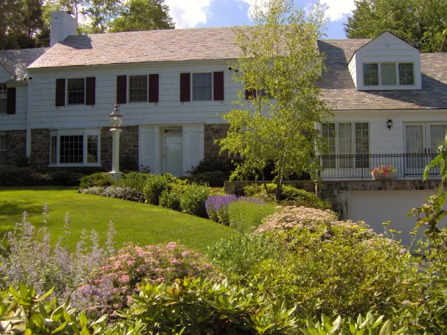 A large house with white siding, stone accents, and a shingled roof, surrounded by a well-maintained garden with green grass, various flowering plants, and a small tree.