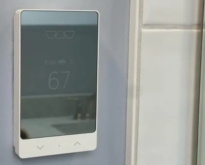 Smart tech bathroom addon showing a sleek white digital touchscreen thermostat controller mounted on a wall next to white tile.