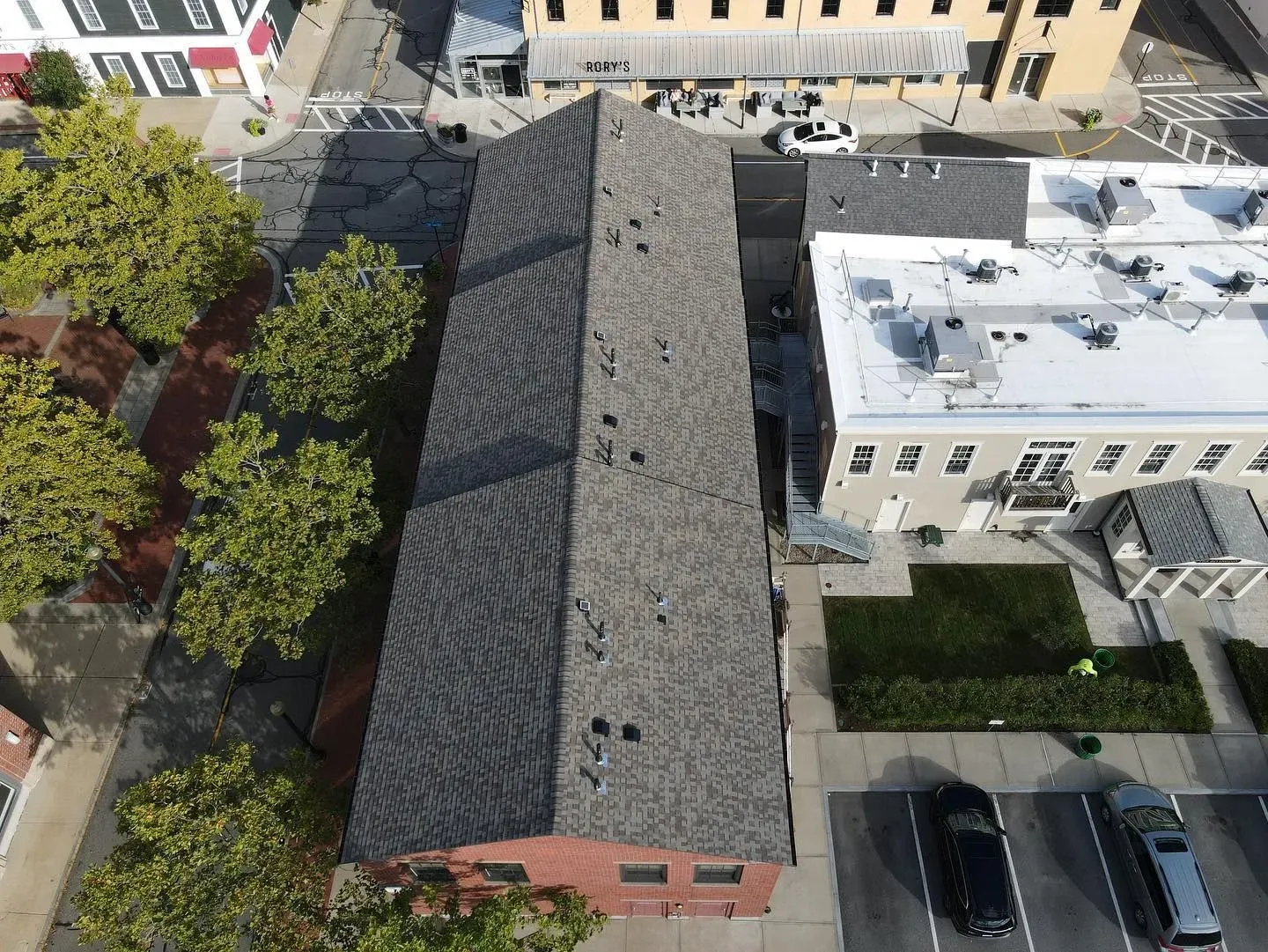 Aerial view of a long gray-roofed building between a street and parked cars in an urban neighborhood