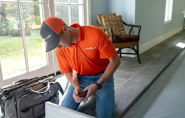 A person in an orange shirt and cap kneels indoors near a tool bag, working with tools beside a window and a wicker chair.
