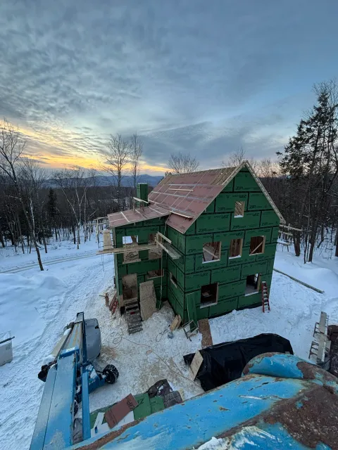 A house under construction on a snowy landscape at sunset, with exposed framing and green exterior sheathing, surrounded by construction tools and materials.