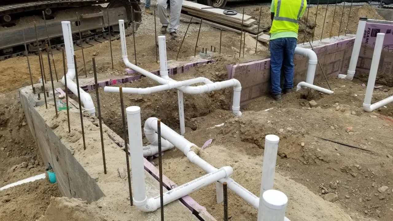 Construction site with exposed white PVC plumbing pipes in the ground, surrounded by dirt and rebar, while two workers in safety gear stand nearby—typical of ongoing projects across Norfolk and Suffolk County.