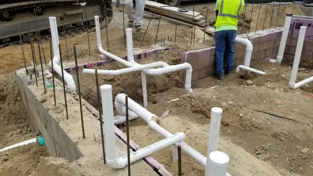 Construction site with exposed white PVC plumbing pipes in the ground, surrounded by dirt and rebar, while two workers in safety gear stand nearby—typical of ongoing projects across Norfolk and Suffolk County.