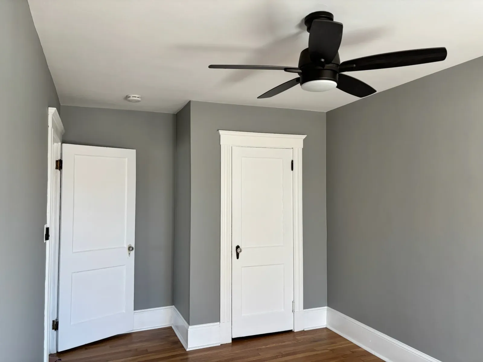 Empty room with gray walls, wood floor, white door, and ceiling fan.