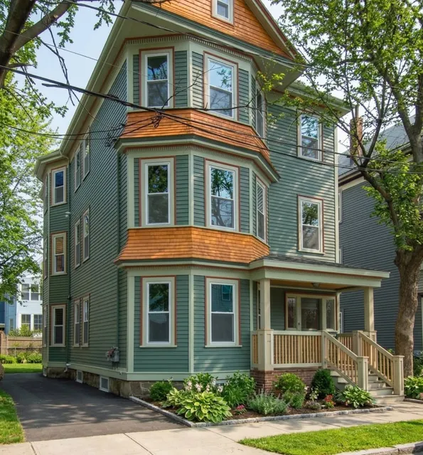 A three-story green house at 17 Smith Place features orange shingle roof accents, large bay windows, a covered porch, and driveway, all surrounded by trees and landscaping.