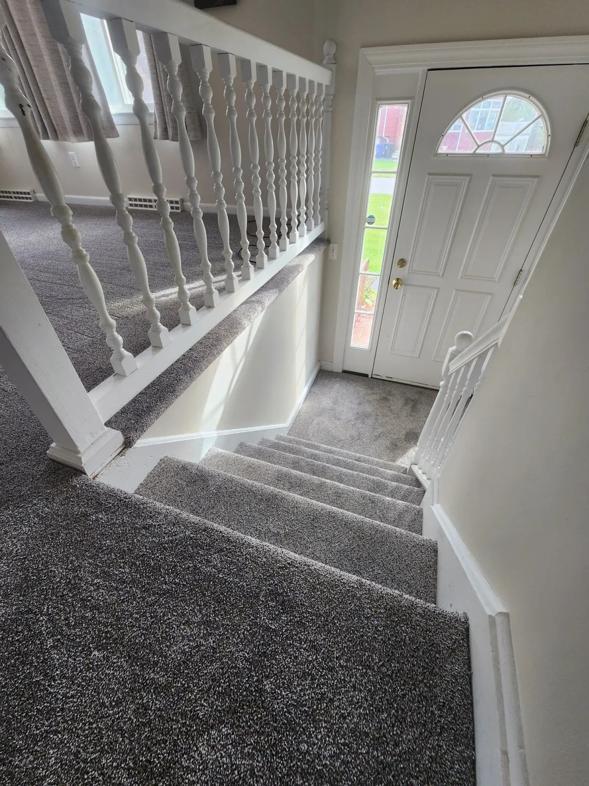 View of a staircase leading to the front door of a house, with white railings, gray carpeted stairs, and a view of the outside through the front door's glass panel.