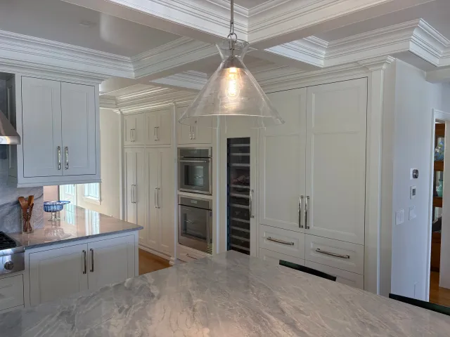 Modern kitchen with white cabinetry and marble countertops, featuring built-in ovens and a glass pendant light