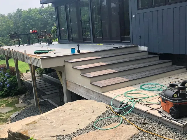 Deck with steps under construction; gray siding; green trees.