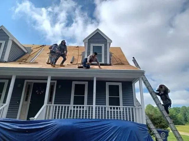Roofers working on a blue-sided house under a sunny sky; one worker climbs a ladder.