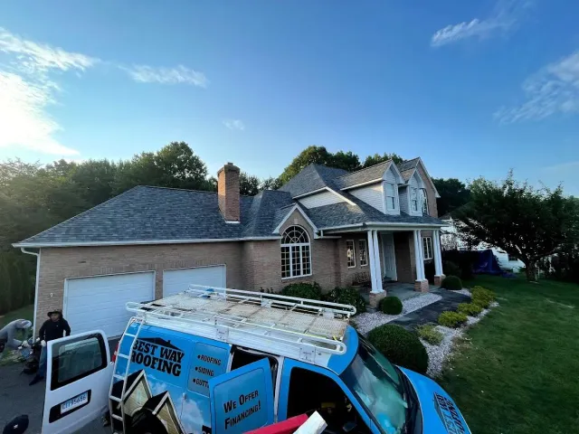 A blue van with a roof rack parked near a brick house with a new roof. A person is near the van.