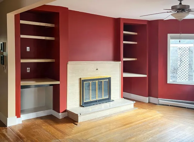 Living room with red walls, a fireplace with light stone surround, built-in wooden shelves, and a ceiling fan, by LSC Carpentry LLC.