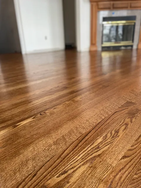 Close-up of a polished wooden floor with visible wood grain, in front of a fireplace with a brick and wood mantel.