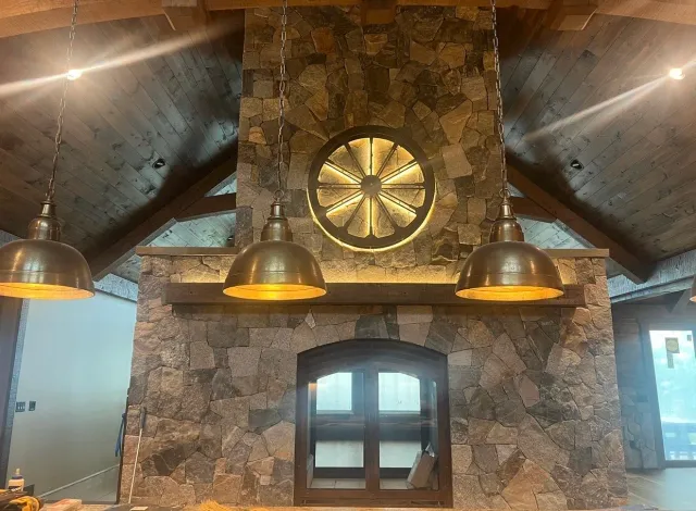 Interior of a house with a stone fireplace, a large round light fixture resembling a wheel, and three hanging metallic pendant lights.