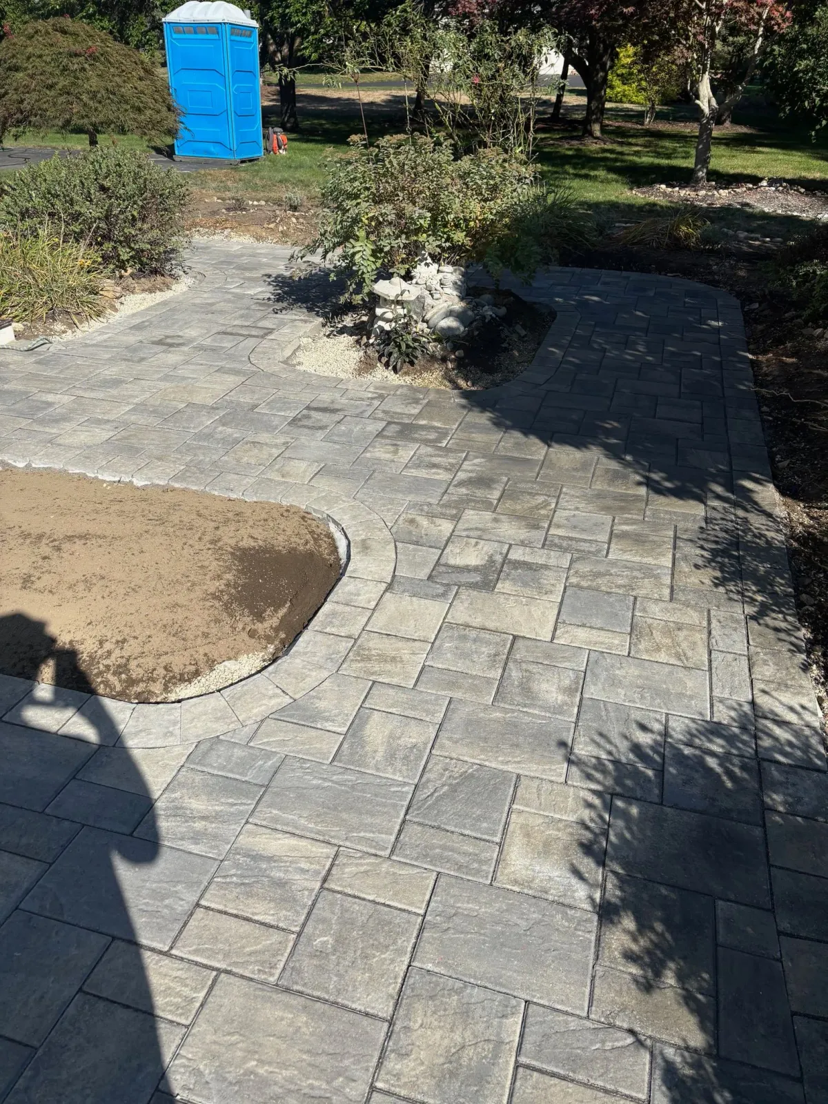 Brick paved pathway in a garden with plants. A blue portable toilet is in the background.