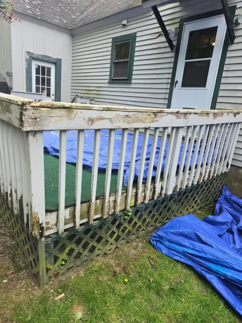 Deteriorating deck railing on residential home with peeling paint, rotted wood beam, white spindles, and blue tarps on ground