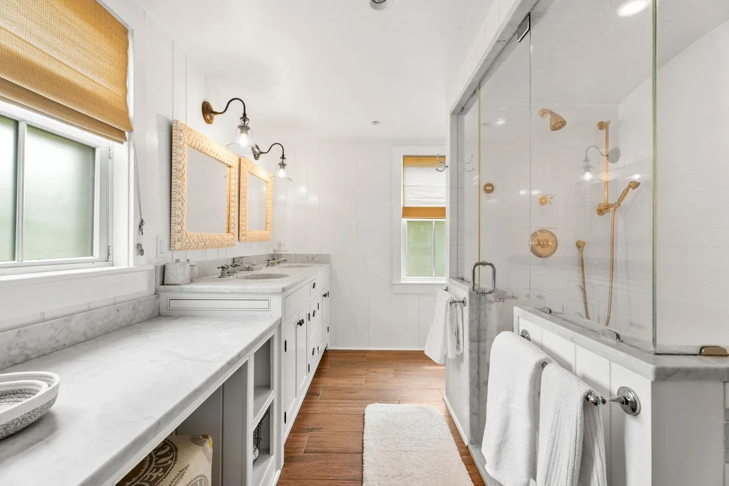 Bright white bathroom with a double vanity, marble countertops, and two framed mirrors. There are wall-mounted lights above the mirrors and a window with a wooden shade. To the right, there is a glass-enclosed shower with gold fixtures, white tiled walls, and wooden flooring outside.