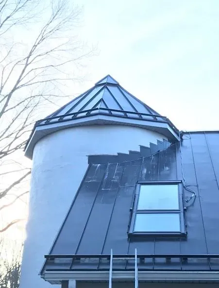 Round tower with gray conical roof beside a dark metal sloped roof and dormer window
