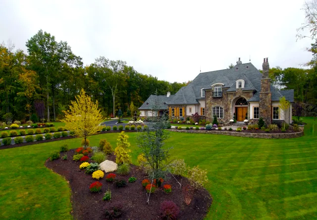 Large house with stone and white exterior, surrounded by a landscaped lawn and garden with various shrubs, trees, and flowers. The background has a wooded area.