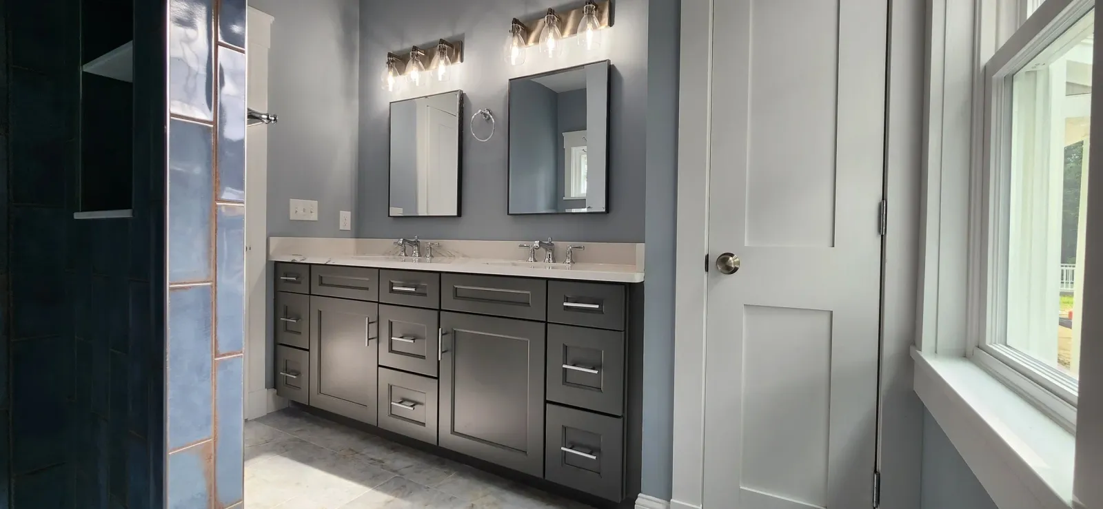 Modern bathroom with double vanity, two mirrors, gray cabinets, and overhead lighting. The walls are painted in a muted gray-blue tone, and the floor is tiled. A window and a wooden door are visible on the right.
