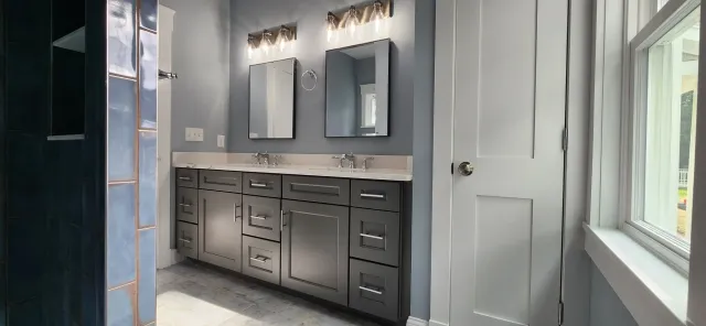 Modern bathroom with double vanity, two mirrors, gray cabinets, and overhead lighting. The walls are painted in a muted gray-blue tone, and the floor is tiled. A window and a wooden door are visible on the right.