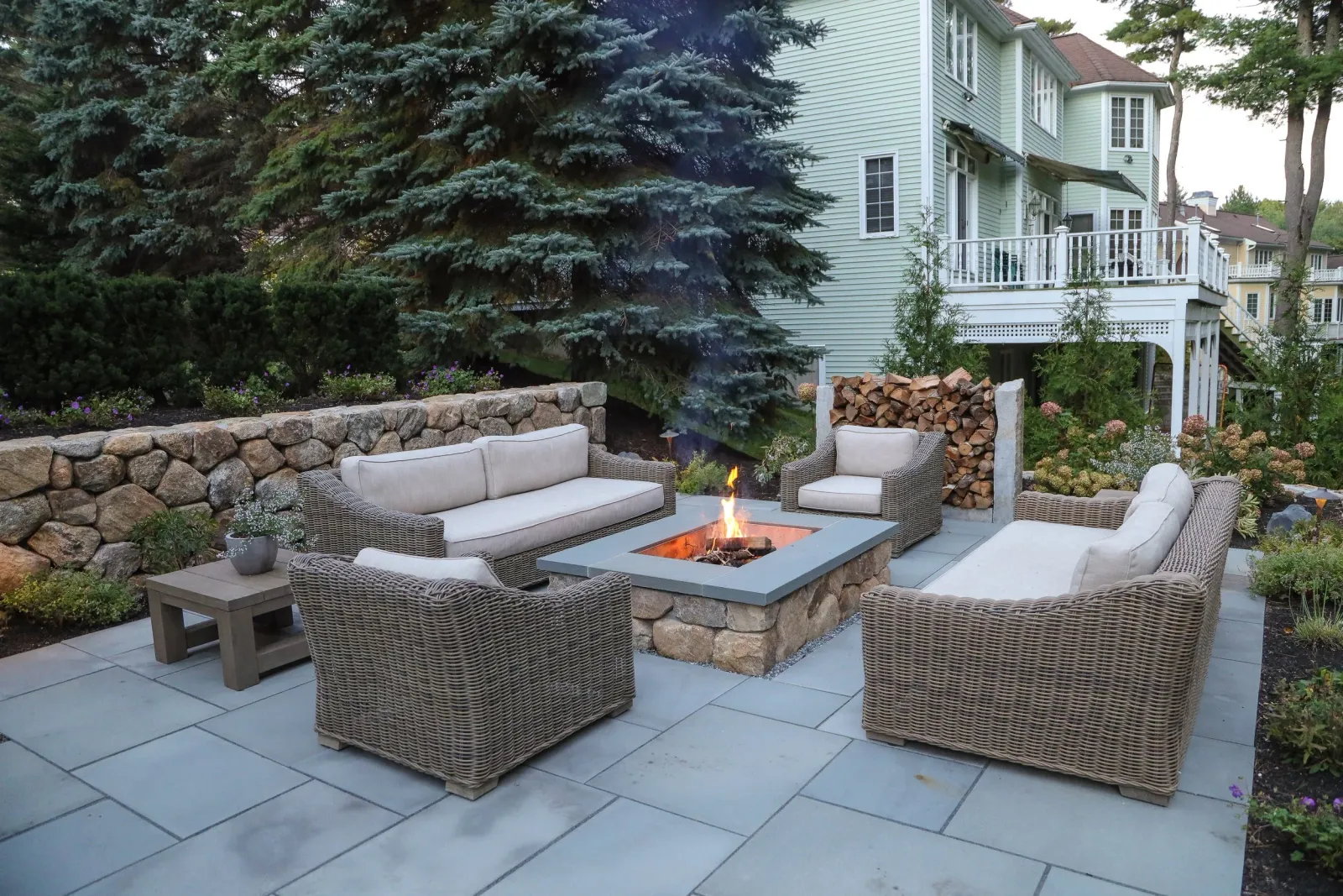 Outdoor patio with wicker furniture and a fire pit, surrounded by garden bushes, trees, and a background of a light green house with a deck.