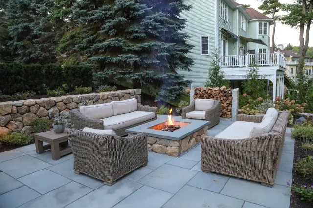 Outdoor patio with wicker furniture and a fire pit, surrounded by garden bushes, trees, and a background of a light green house with a deck.