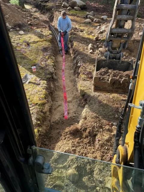 An excavation specialist stands in a waterline trench measuring a pink survey tape while operating a small excavator outside in a grassy and rocky area.