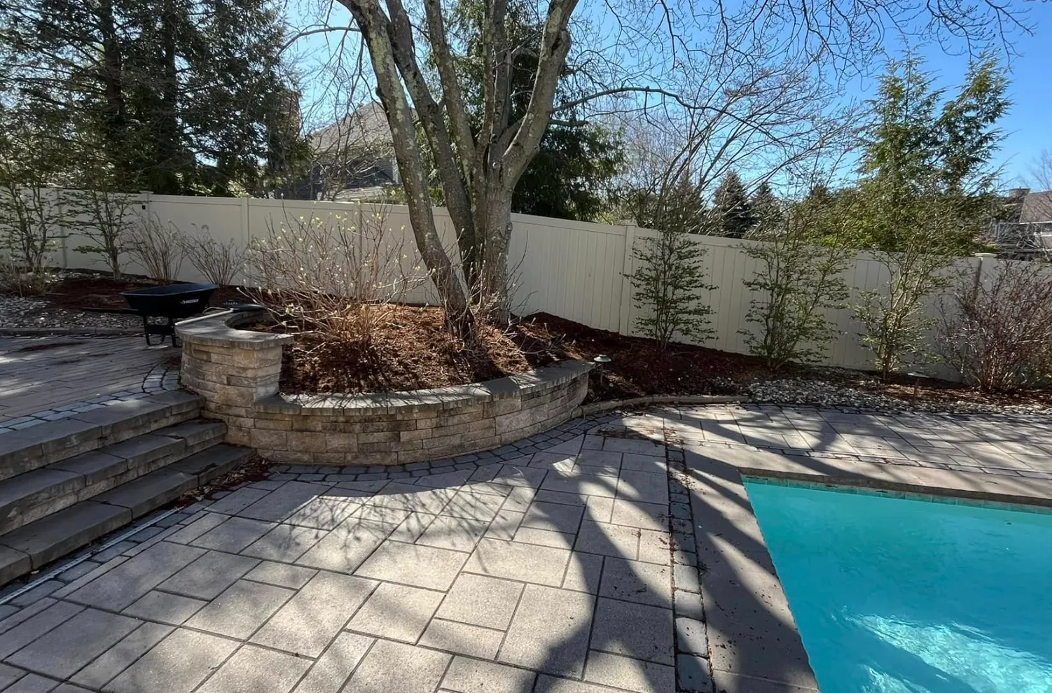 Backyard patio with a swimming pool, brick and stone steps, a tree, surrounding shrubbery, and a white fence under a clear blue sky.