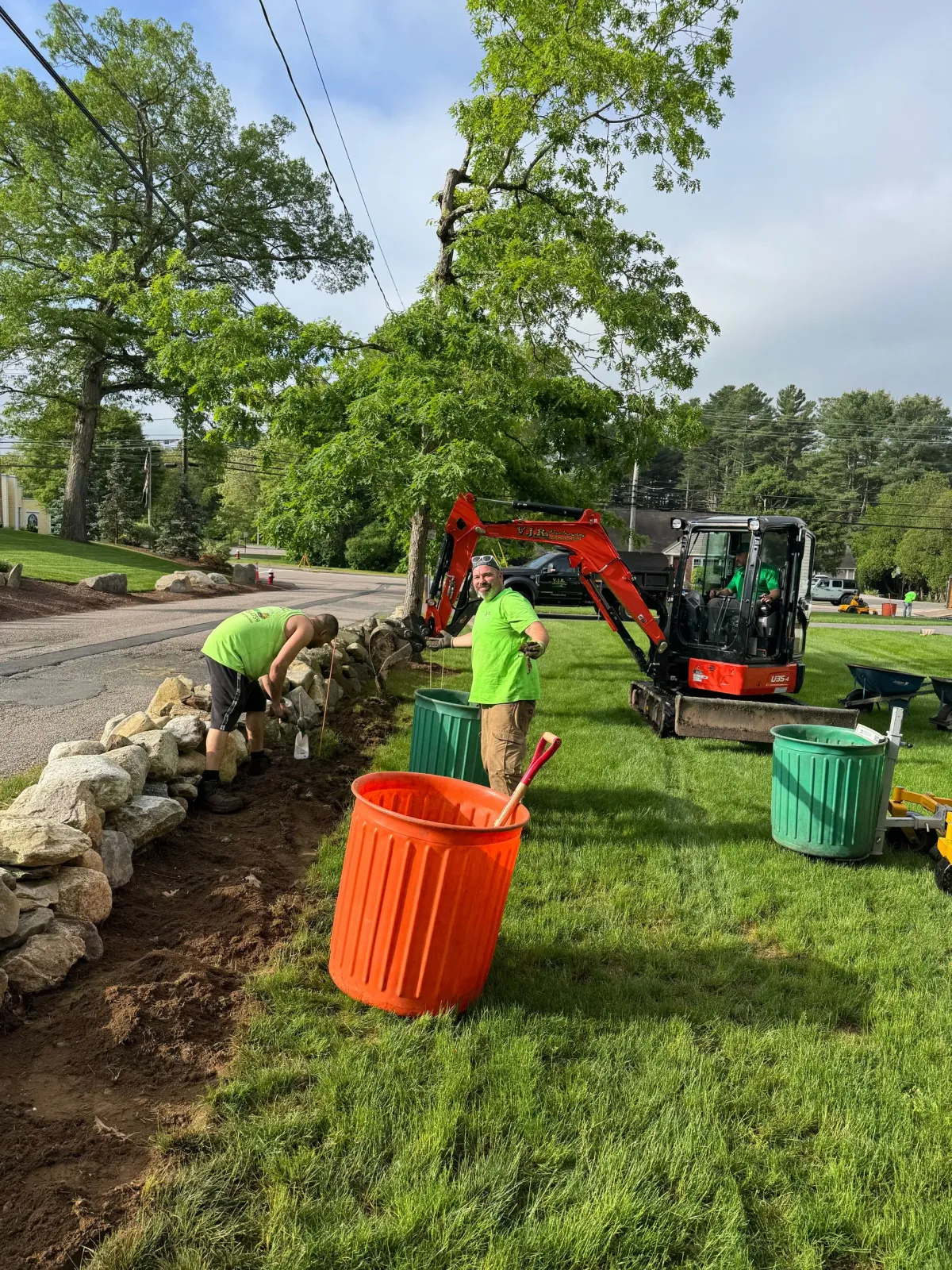 Three people working on landscaping near a stone wall, using an excavator and trash cans on a lawn.