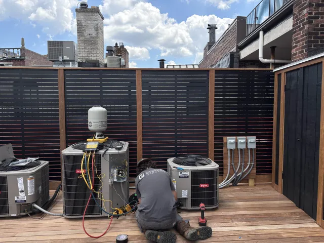 Technician working on rooftop air conditioning units with tools and instruments, surrounded by wood decking and privacy fence.